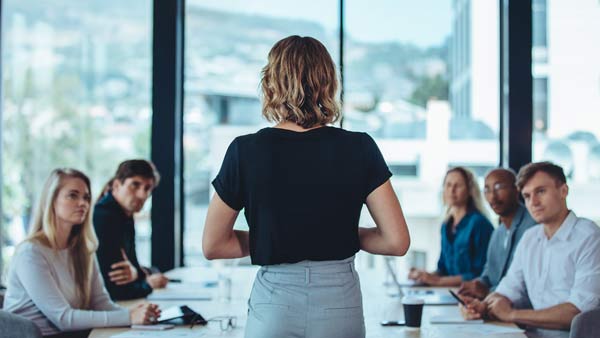 Woman standing at the head of a boardroom table at which 5 people are sat